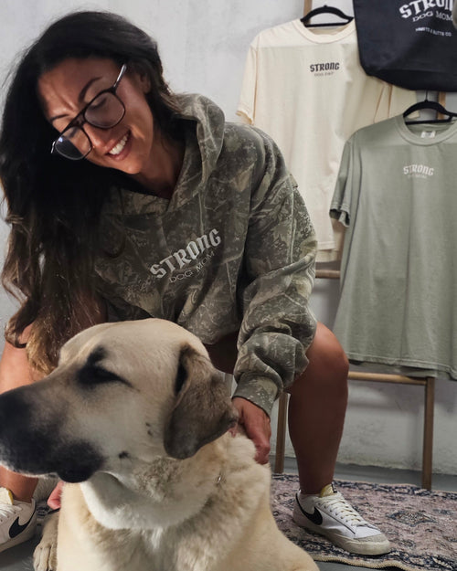 A woman wearing a camouflage hoodie and glasses pets a large cream-colored dog sitting on a table, with clothing racks in the background displaying t-shirts and a tote bag.
